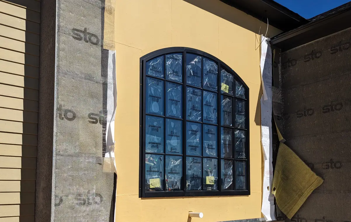 New construction featuring a large arched window with black trim, surrounded by yellow exterior siding and building materials on the ground